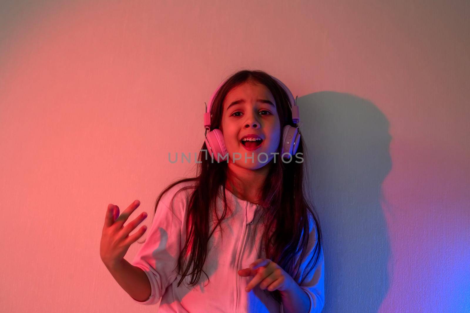 Dancing and singing little girl in pink headphones with loose dark hair in neon pink and blue light near the wall