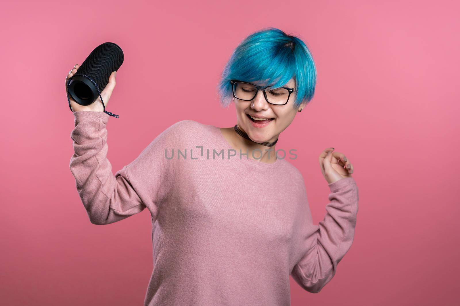 Cute woman with blue hair smiling and dancing with wireless portable speaker in pink studio background. Music, dance concept.