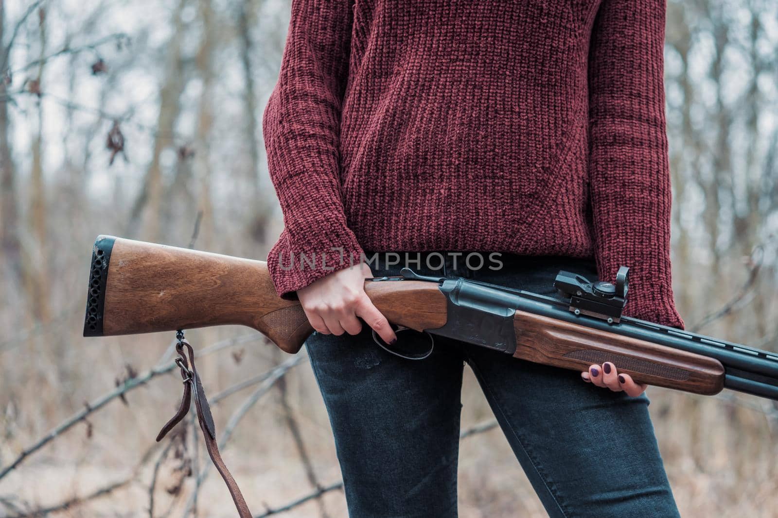 Hunter woman in burgundy warm clothes with gun. Girl hunting in the forest holding her rifle close up. High quality photo