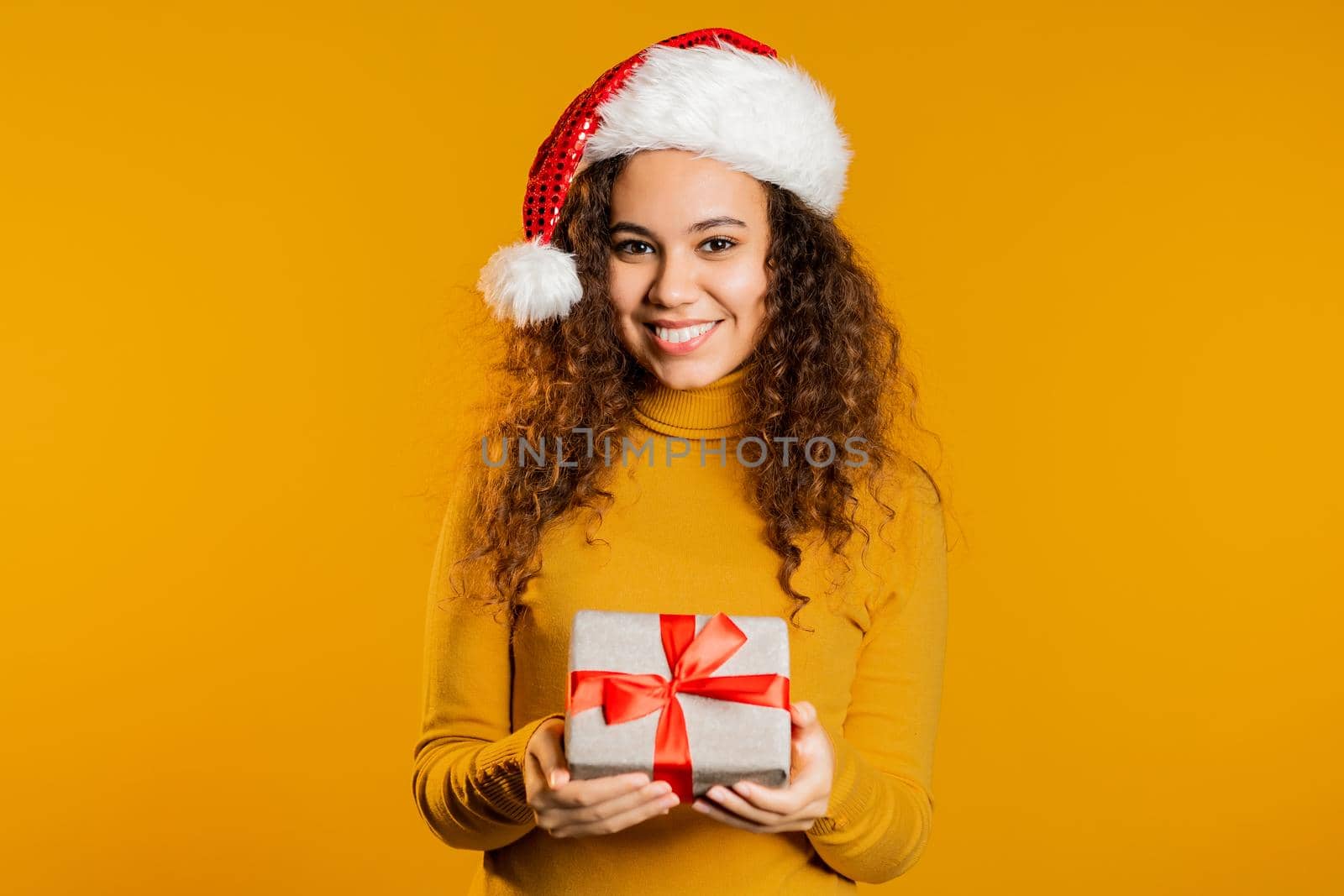 Excited woman in Santa hat received gift box with bow. She is happy and flattered by attention. Girl with present on yellow background. Studio shot by kristina_kokhanova