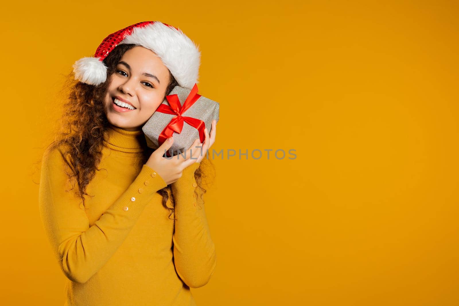 Excited woman in Santa hat received gift box with bow. She is happy and flattered by attention. Girl with present on yellow background. Studio shot. High quality photo