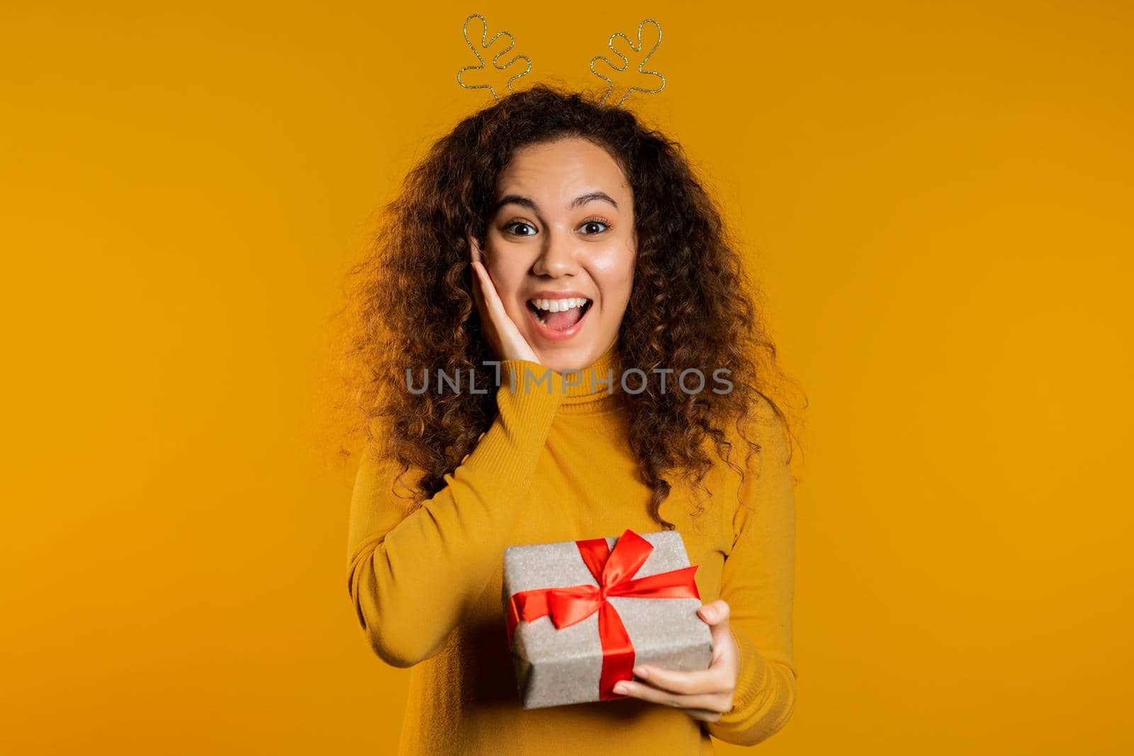 urprised mixed race woman smiling and holding gift box on yellow studio background. Girl with curly hairstyle in sweater. Christmas mood. by kristina_kokhanova