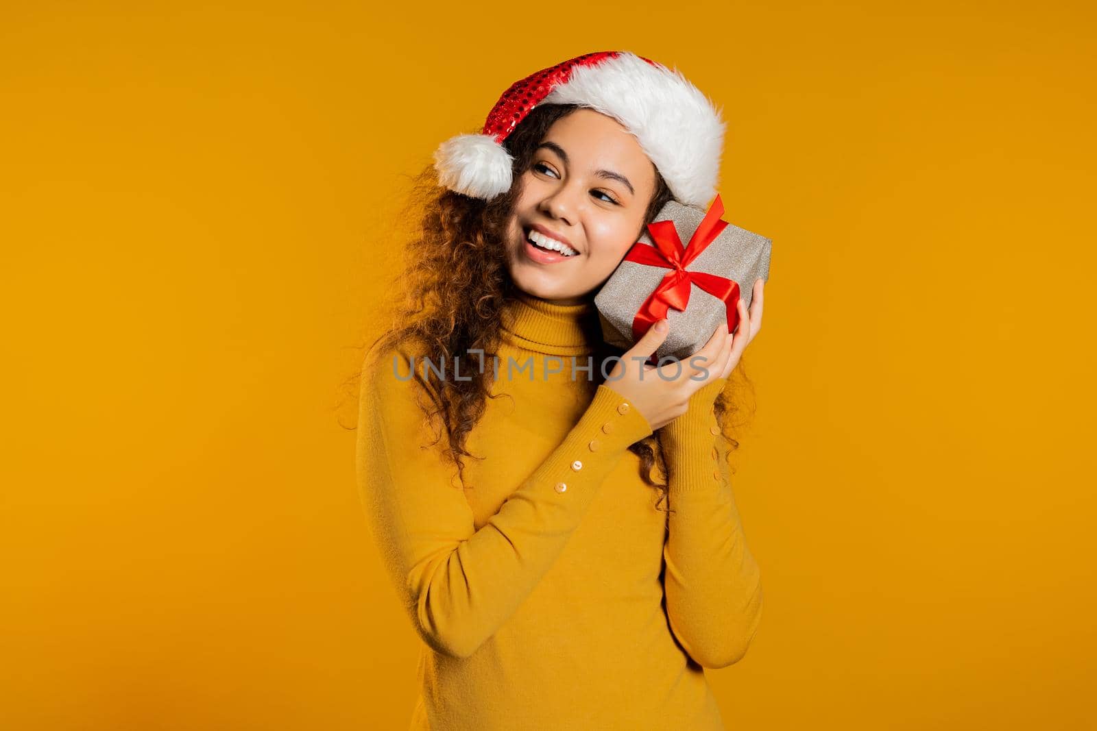 Excited woman in Santa hat received gift box with bow. She is happy and flattered by attention. Girl with present on yellow background. Studio shot. High quality photo