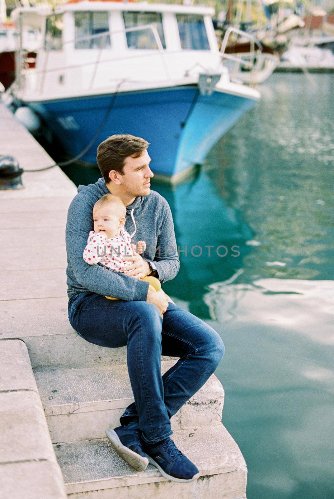 Dad with a baby on his knees sits on the pier near the boat and looks at the sea by Nadtochiy