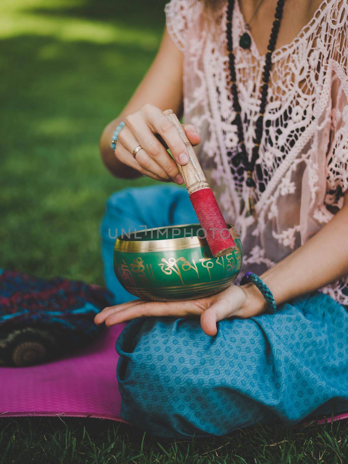 Woman playing on singing bowl while sitting on yoga mat in green park at summer. Vintage tonned. Beautiful girl with mala beads meditating on green grass. by kristina_kokhanova