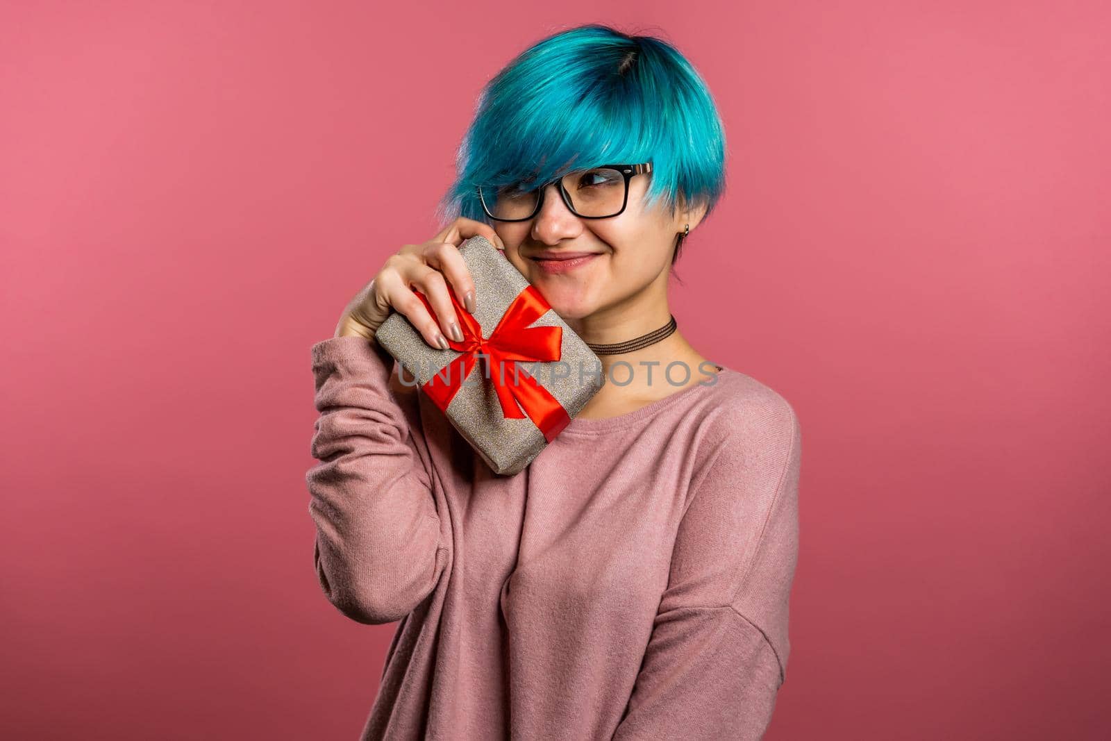 Young unusual woman with blue hair smiling and holding gift box on pink studio background. Girl cozy sweater and glasses. Christmas mood. by kristina_kokhanova