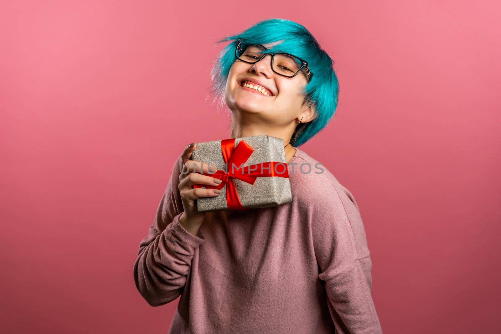 Young unusual woman with blue hair smiling and holding gift box on pink studio background. Girl cozy sweater and glasses. Christmas mood. by kristina_kokhanova