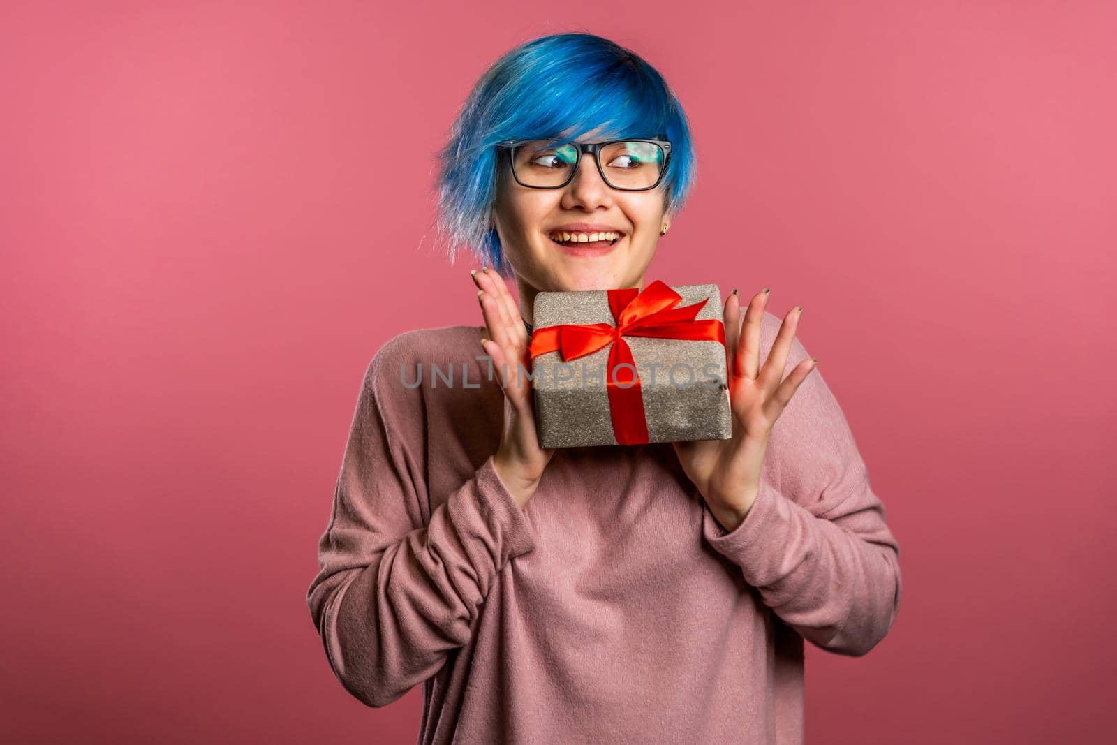 Young unusual woman with blue hair smiling and holding gift box on pink studio background. Girl cozy sweater and glasses. Christmas mood. by kristina_kokhanova