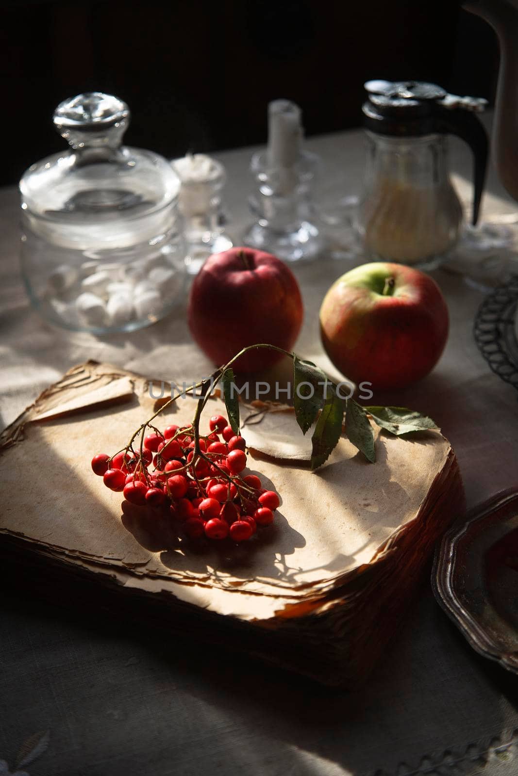 A bunch of red rowan lying on the old and scrubby book, two red apples and the other tableware for tea time on gray rustic table in the morning light, selective focus.