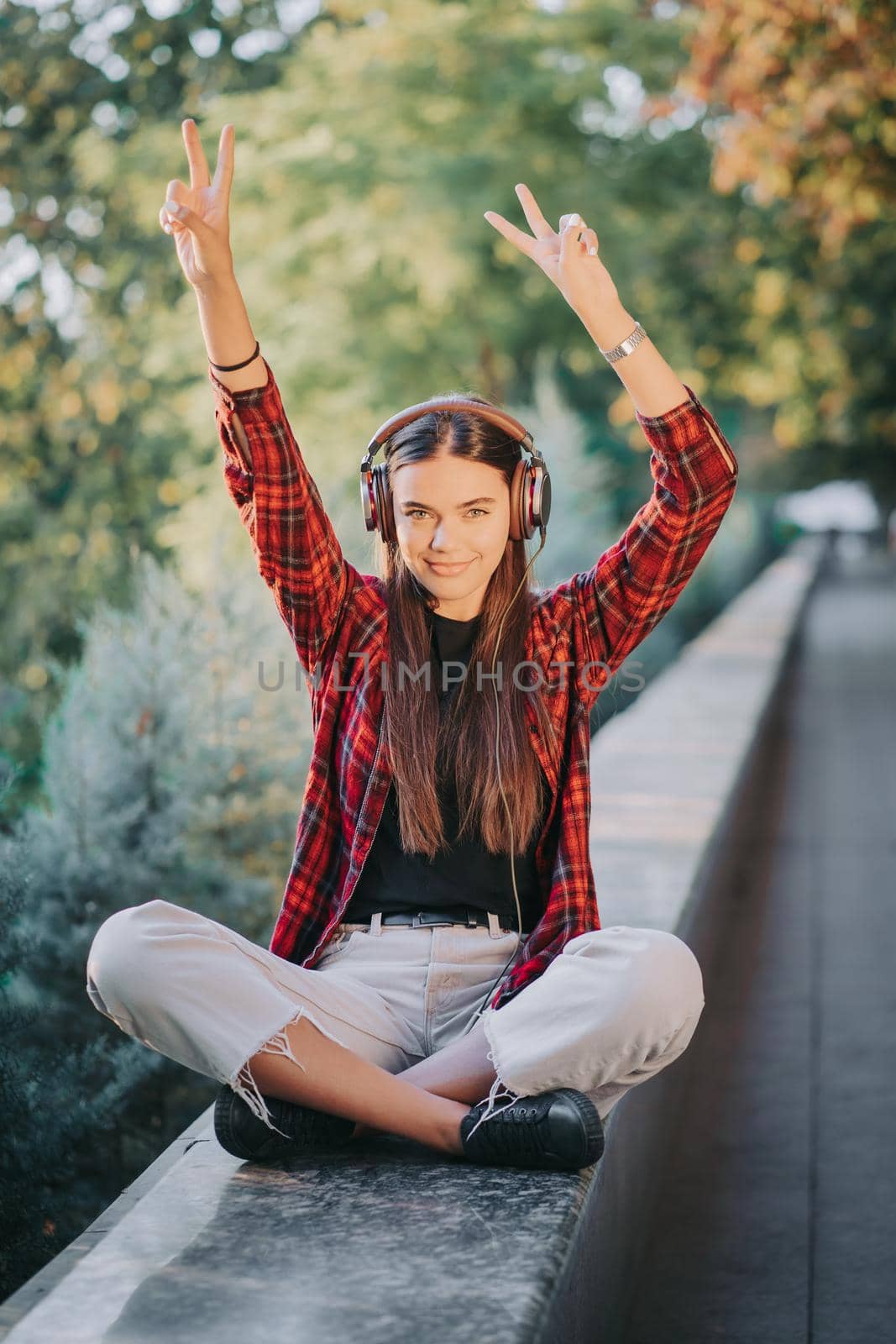 Young teenager listens to music through headphones in park.Girl in red plaid shirt smiles, dancing to rhythm.Concept of student life, freedom, modern youth.