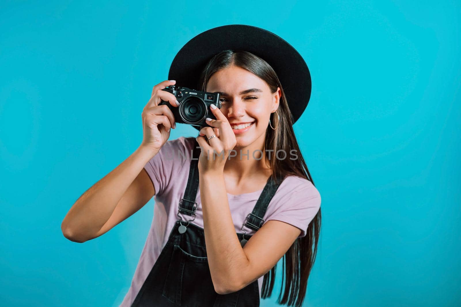 Young pretty woman in overall takes pictures with DSLR camera over blue background in studio. Girl smiling and having fun as photographer. High quality photo