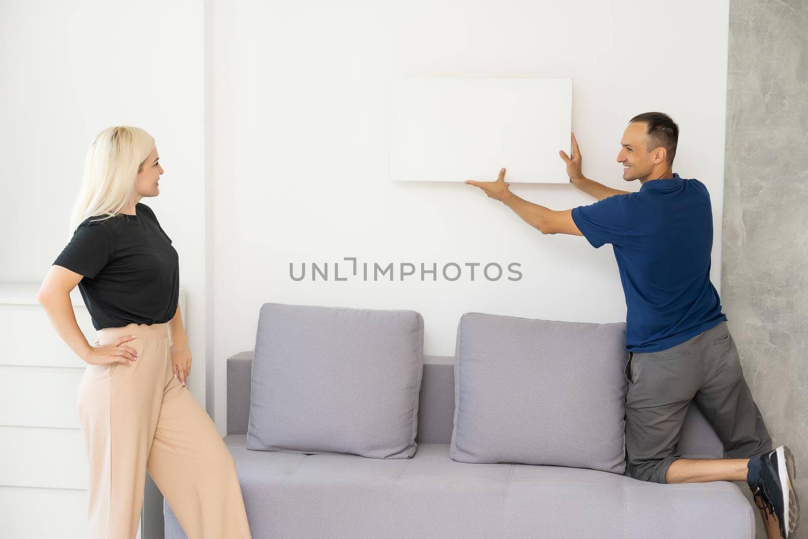 Excited young husband and wife hang frame picture on wall happy settling together moving to new house, overjoyed biracial couple decorating own home relocating, ownership concept.