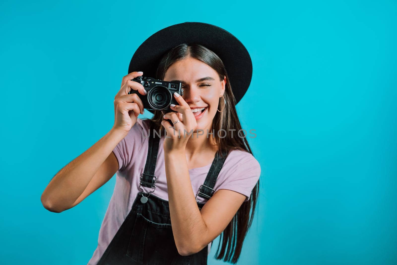 Young pretty woman in overall takes pictures with DSLR camera over blue background in studio. Girl smiling and having fun as photographer. High quality photo