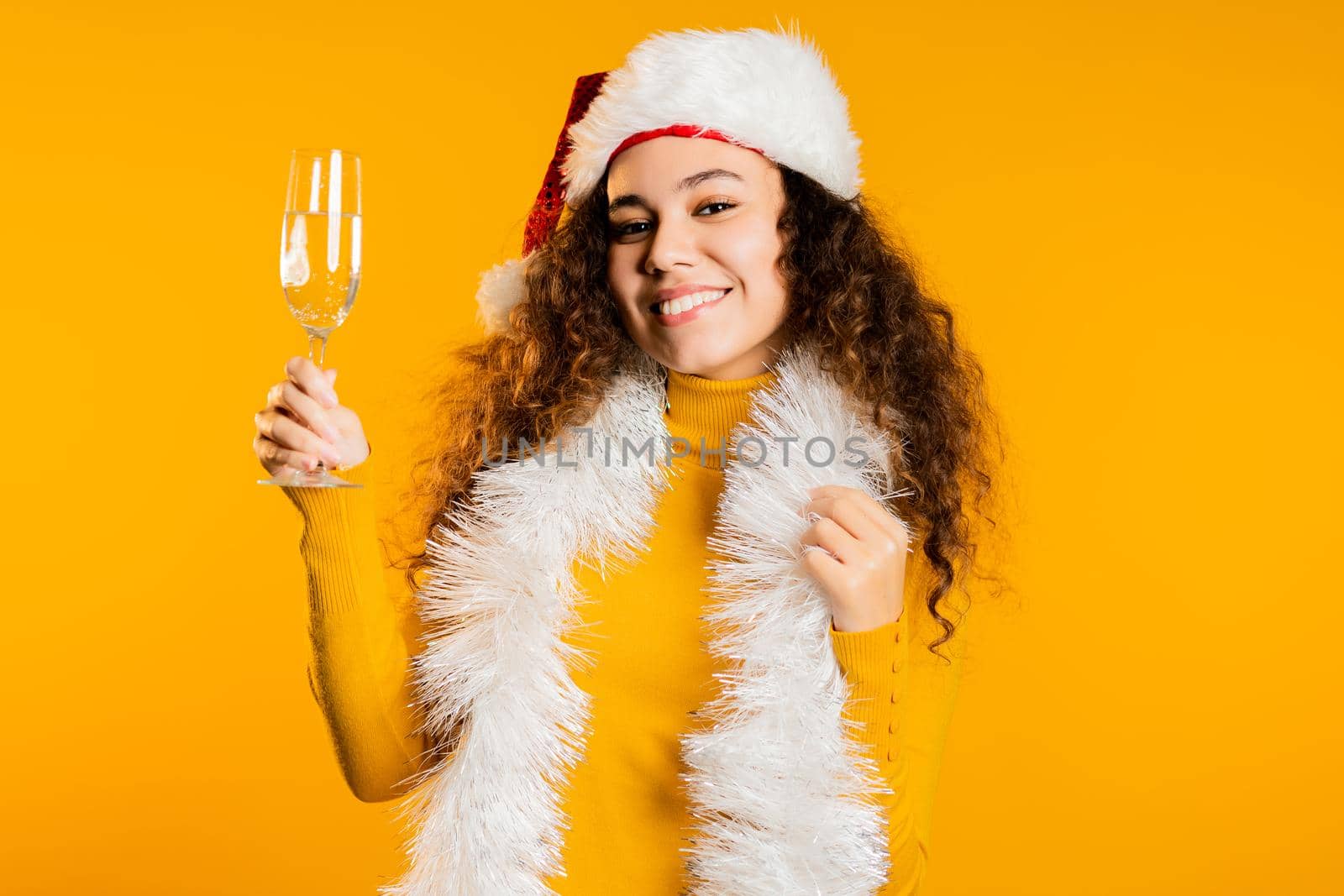 Young cute girl smiling and holding glass of champagne or wine on yellow studio background. Woman in Santa hat. New year mood. High quality photo