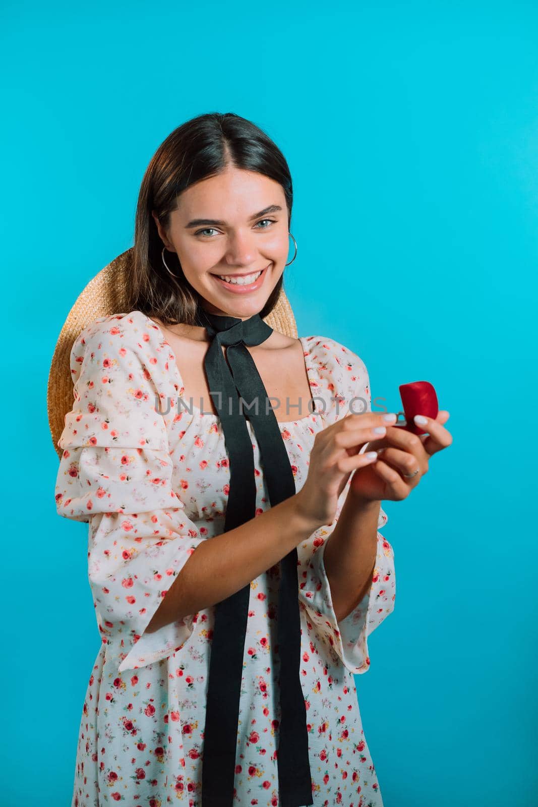 Attractive young woman holding small jewelry box with proposal diamond ring on blue wall background. Lady smiling, she is happy to get present, proposition for marriage.