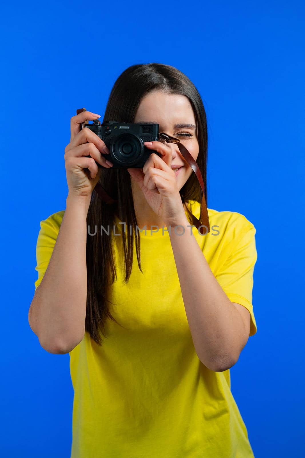 Young pretty brunette woman in yellow shirt takes pictures with DSLR camera over blue background in studio. Girl smiling, having fun as photographer.