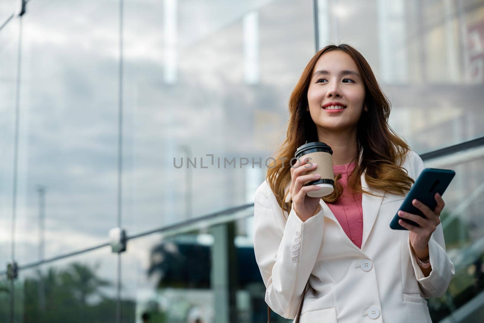 Young business woman smiling holding mobile phone with coffee take away going to work early in morning, Asian businesswoman with smartphone and cup coffee standing against street building near office