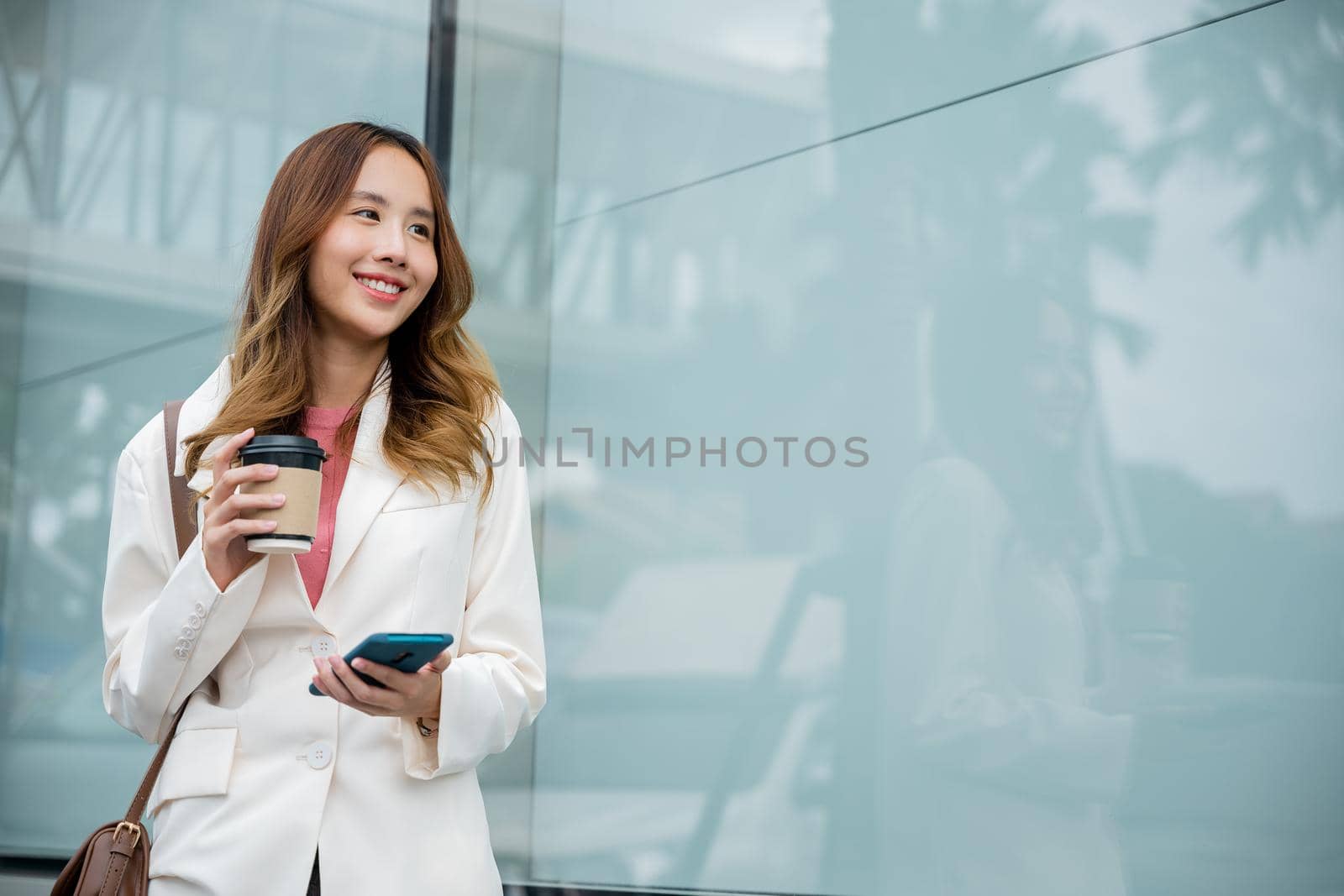 Asian businesswoman with smartphone and cup coffee standing against street building near office by Sorapop