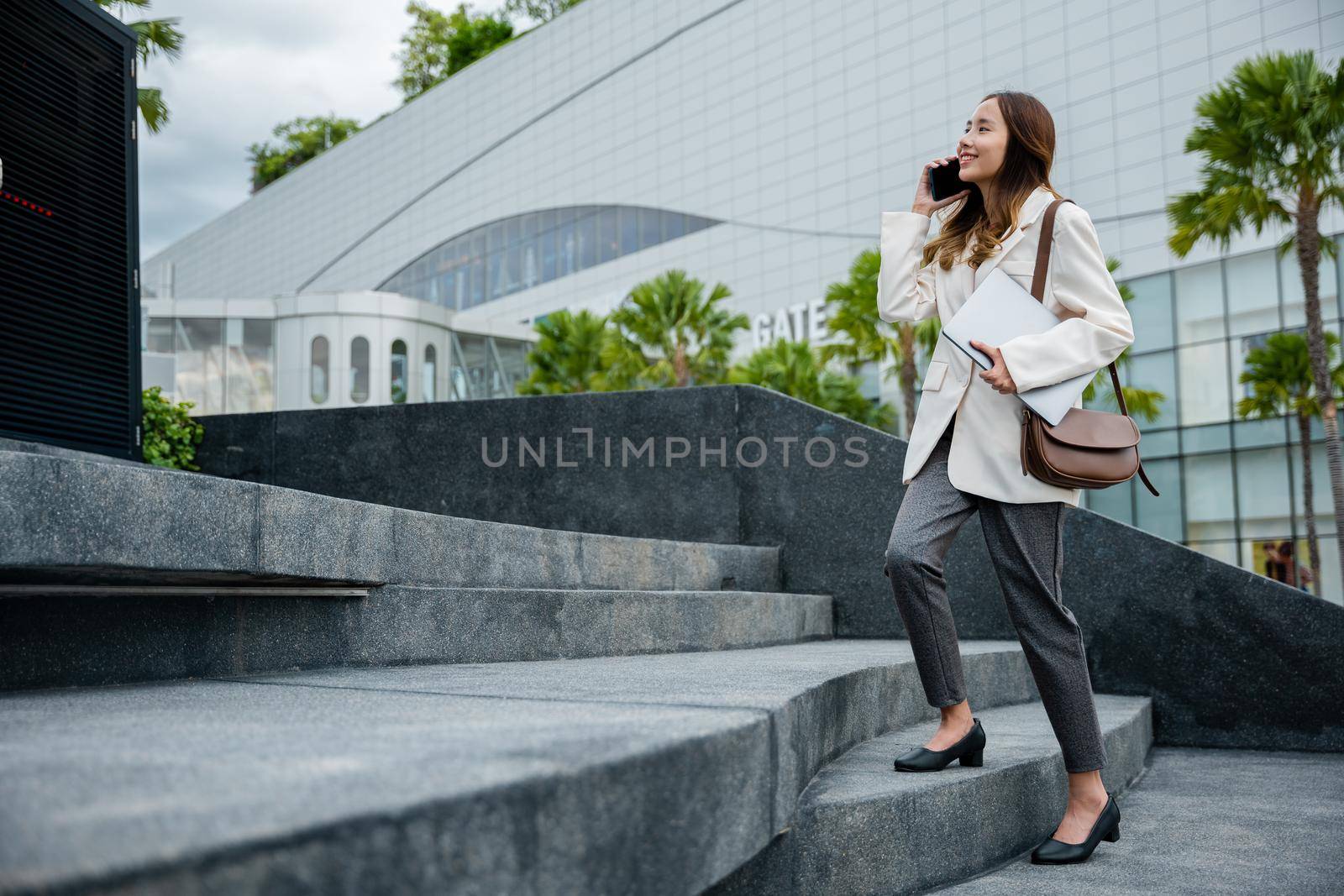 Business woman hold laptop computer talking mobile phone she hurry up walking on stairway by Sorapop
