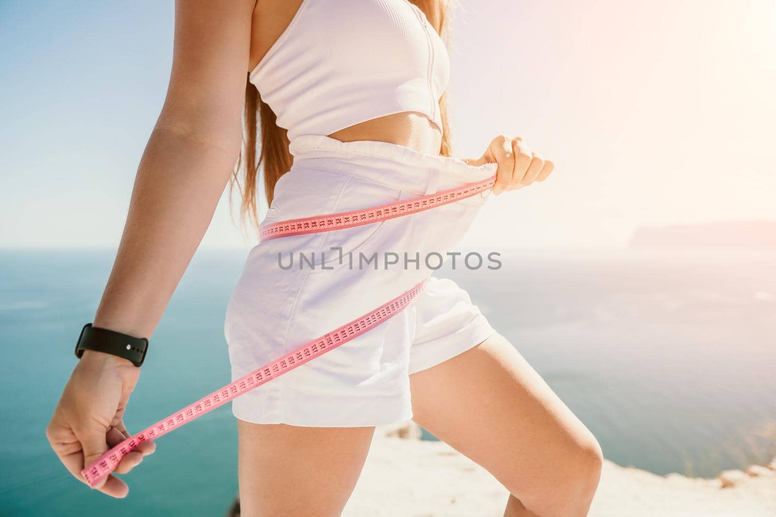 a young and slender woman in shorts holds in her hand a measuring yellow tape on her sexy thighs on the beach. concept of detox and diet
