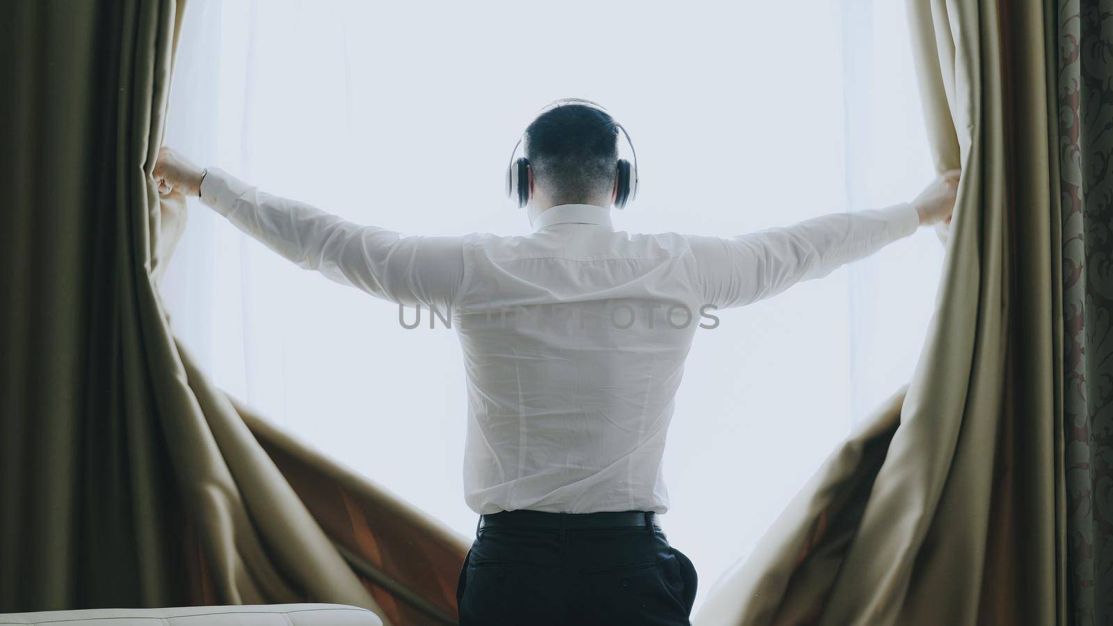 Joyful businessman unveil curtains in hotel room at the morning and dancing listening music with wireless headphones looking into window