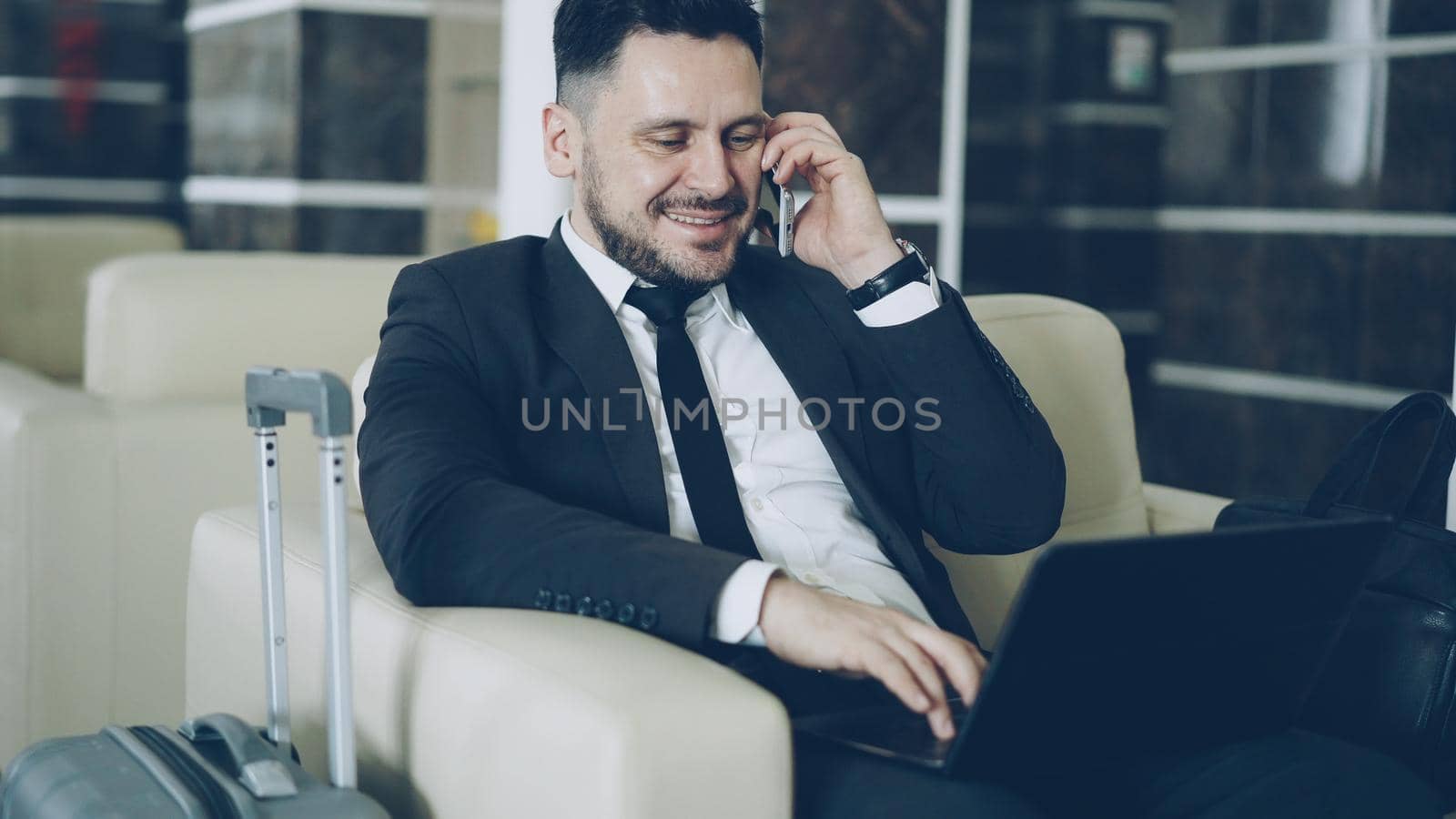 Tilt up of confident attractive businessman talking mobile phone and using laptop computer while sitting on armchair in luxury hotel with luggage suitcase near him. Travel, business and people concept