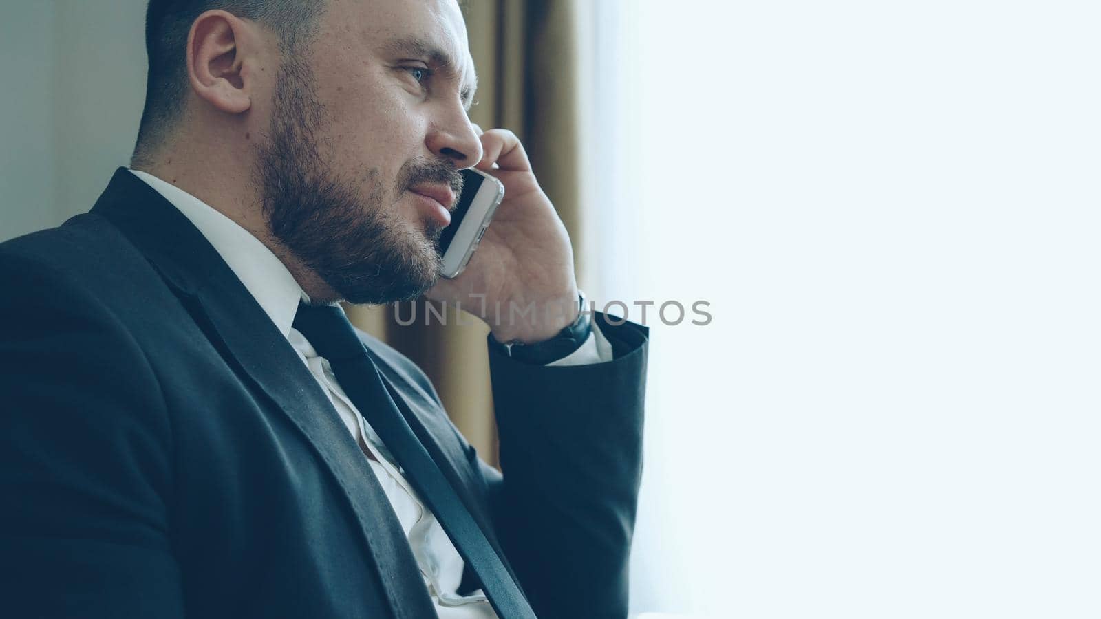 Close-up of confident businessman talking mobile phone and writing notes in notepad while sits on armchair in hotel room or office. Travel, business and people concept by silverkblack