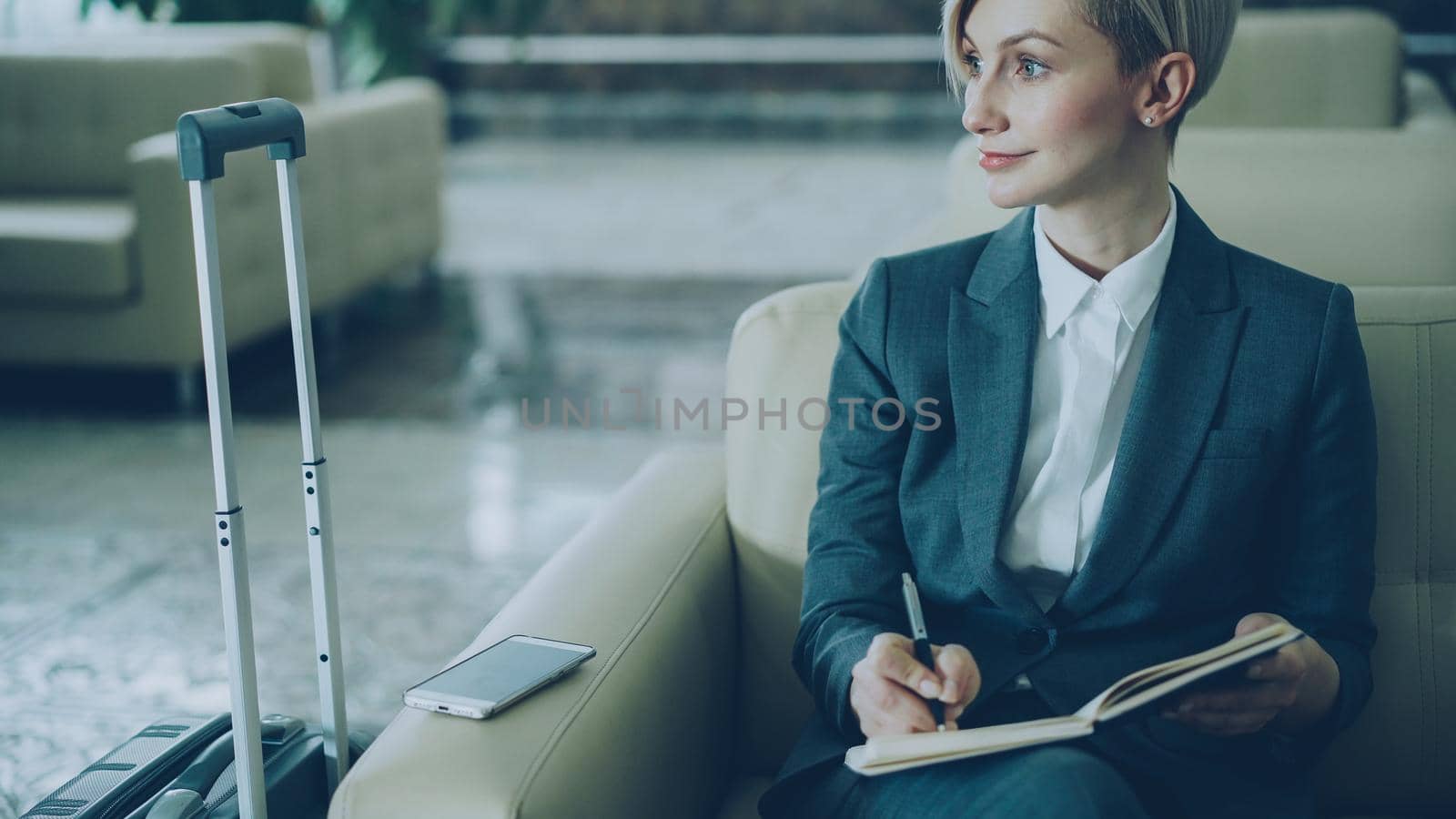 Attractive blonde businesswoman sitting in armchair in hotel lobby and writing in notepad smiling and looking aside. Business, travel and people concept