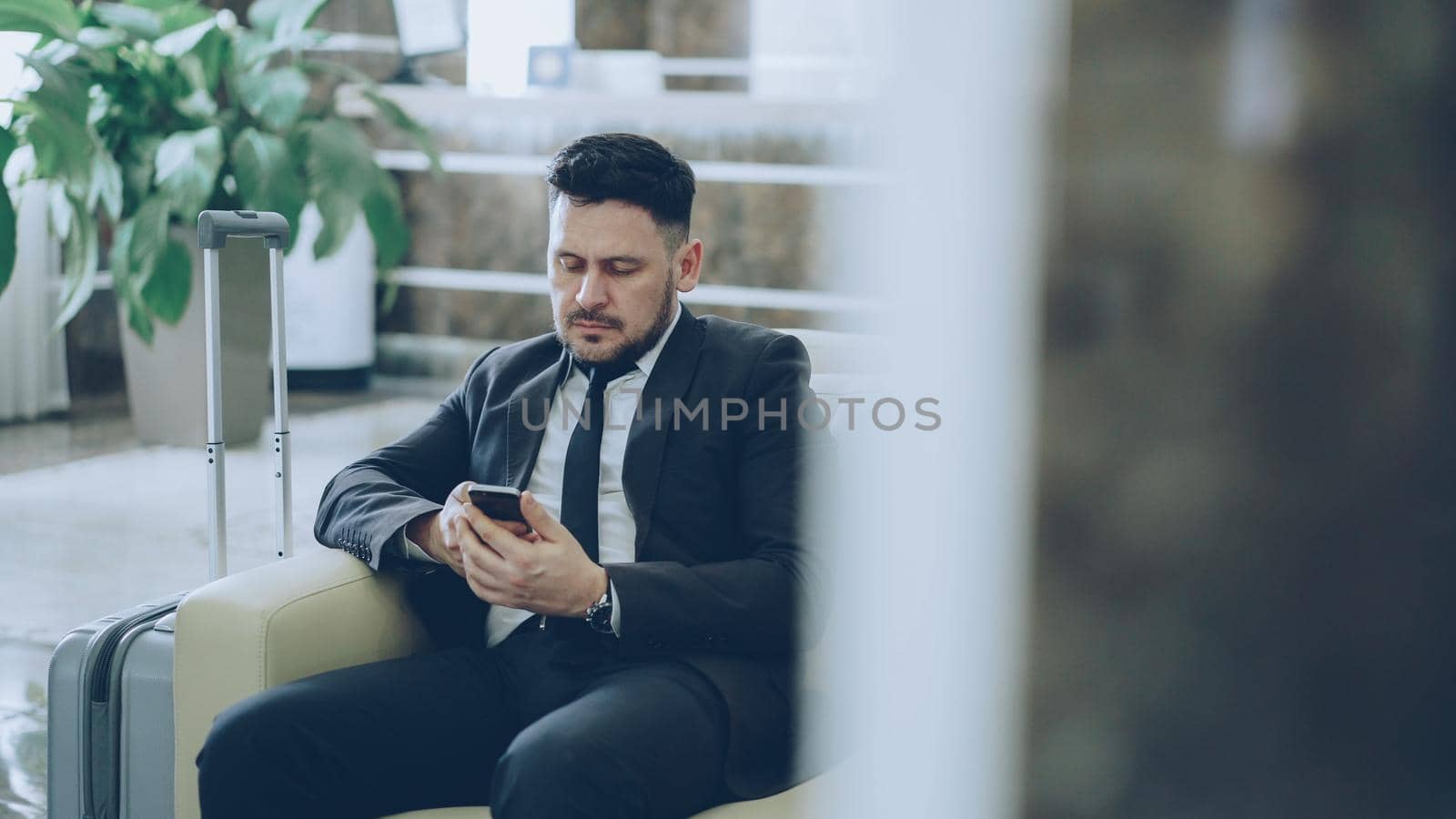 Bearded concentrated businessman using smartphone sit on armchair inside luxury hotel after arrivel to business meeting by silverkblack