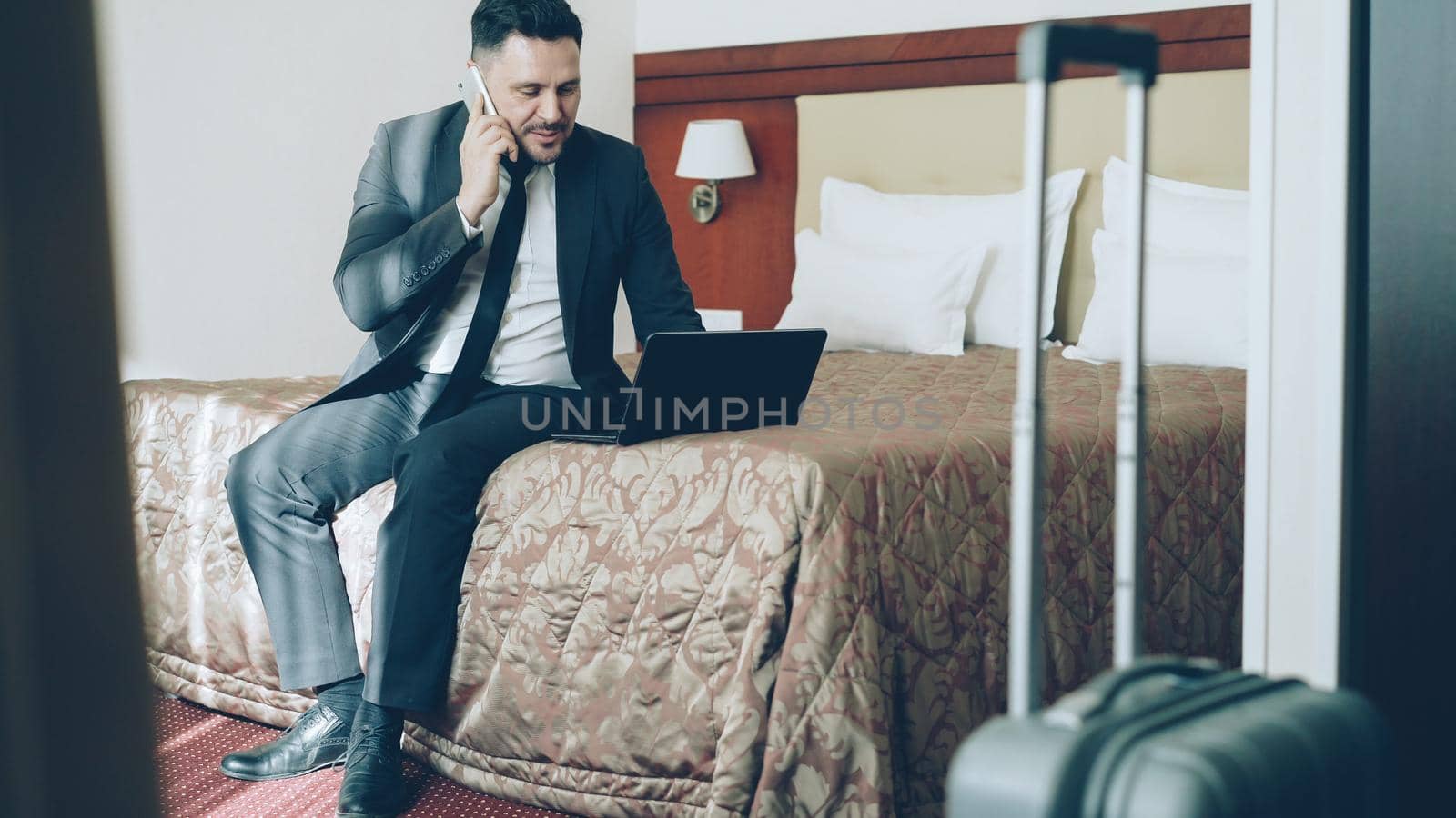 Tilt up of busy young businessman in suit working on laptop and talking at mobile phone while sitting on bed in hotel room. Business, travel and people concept