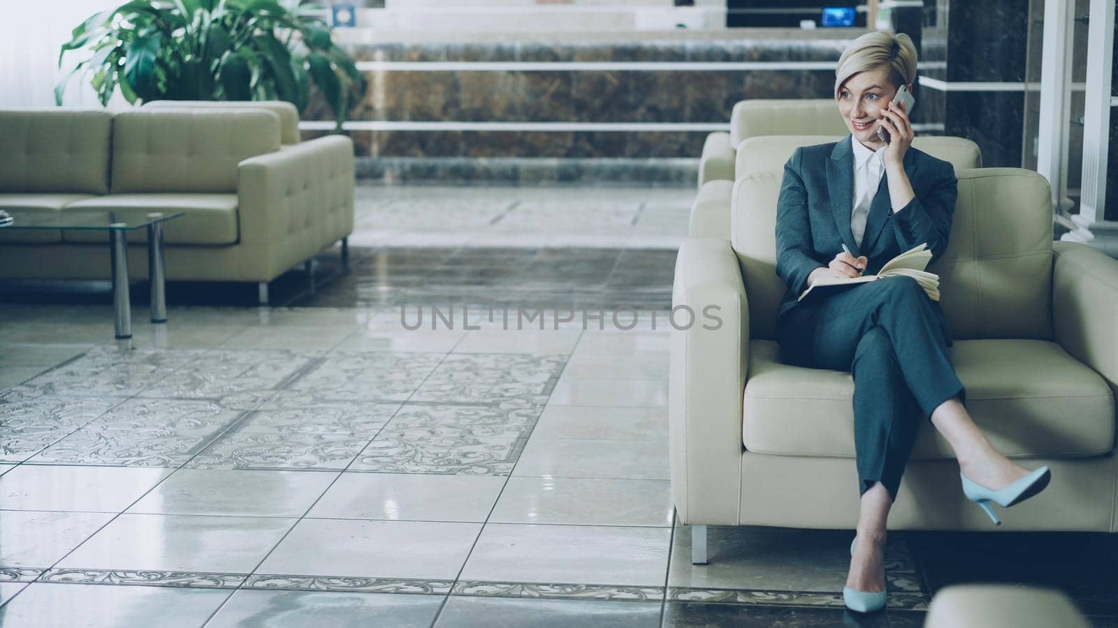 Cheerful blonde businesswoman sitting in armchair in hotel lobby talking on mobile phone and writing in notepad smiling and looking aside. Business, travel and people concept by silverkblack