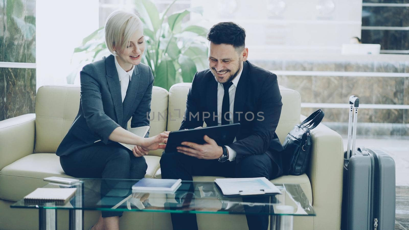 Two female and male business colleagues using digital tablet, notepad and talking at hotel lobby while sitting on couch and waiting check in at reception