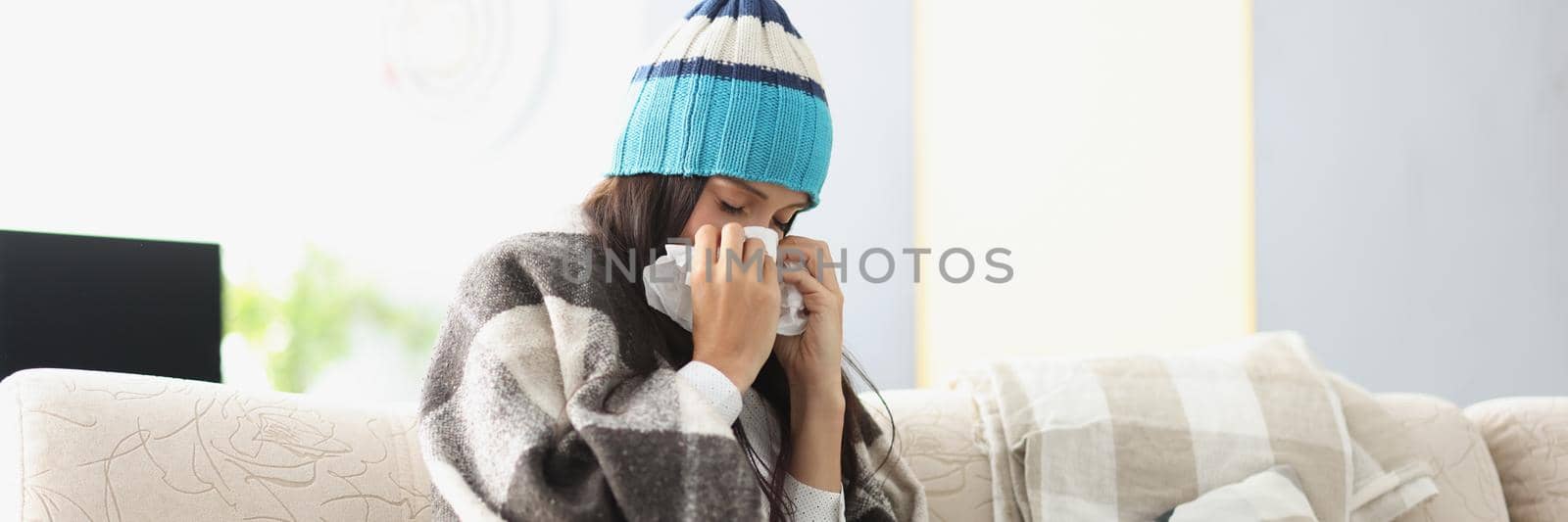 Sick woman in warm hat blowing her nose into paper napkin at home by kuprevich