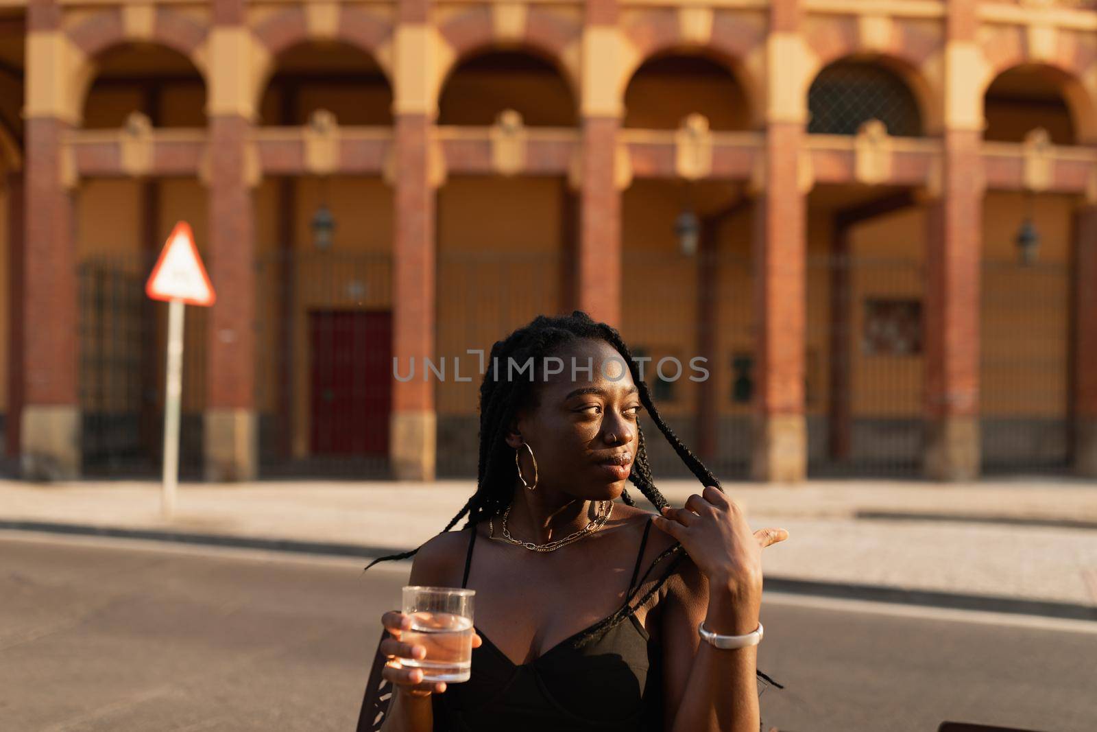 Portrait of a young black female looking at the distance caressing her hair while holding a glass of water at sunset