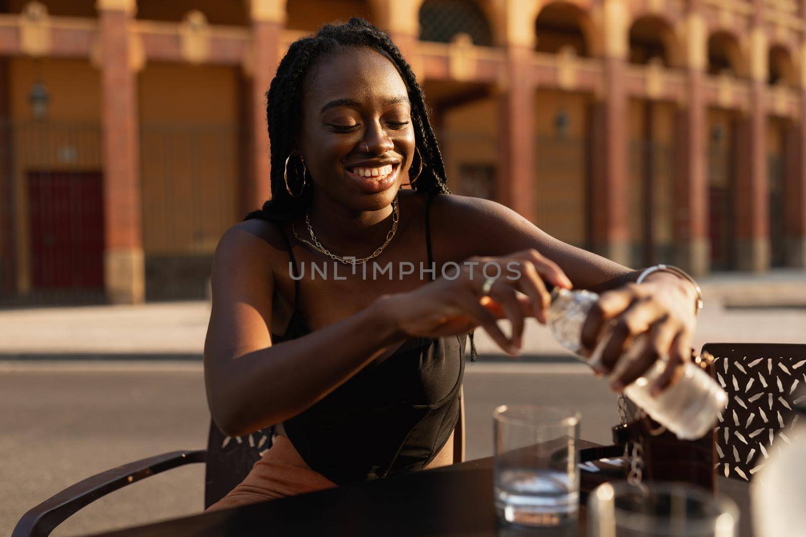 Young black woman closing a bottle of water while resting in a bar terrace in the city during sunset