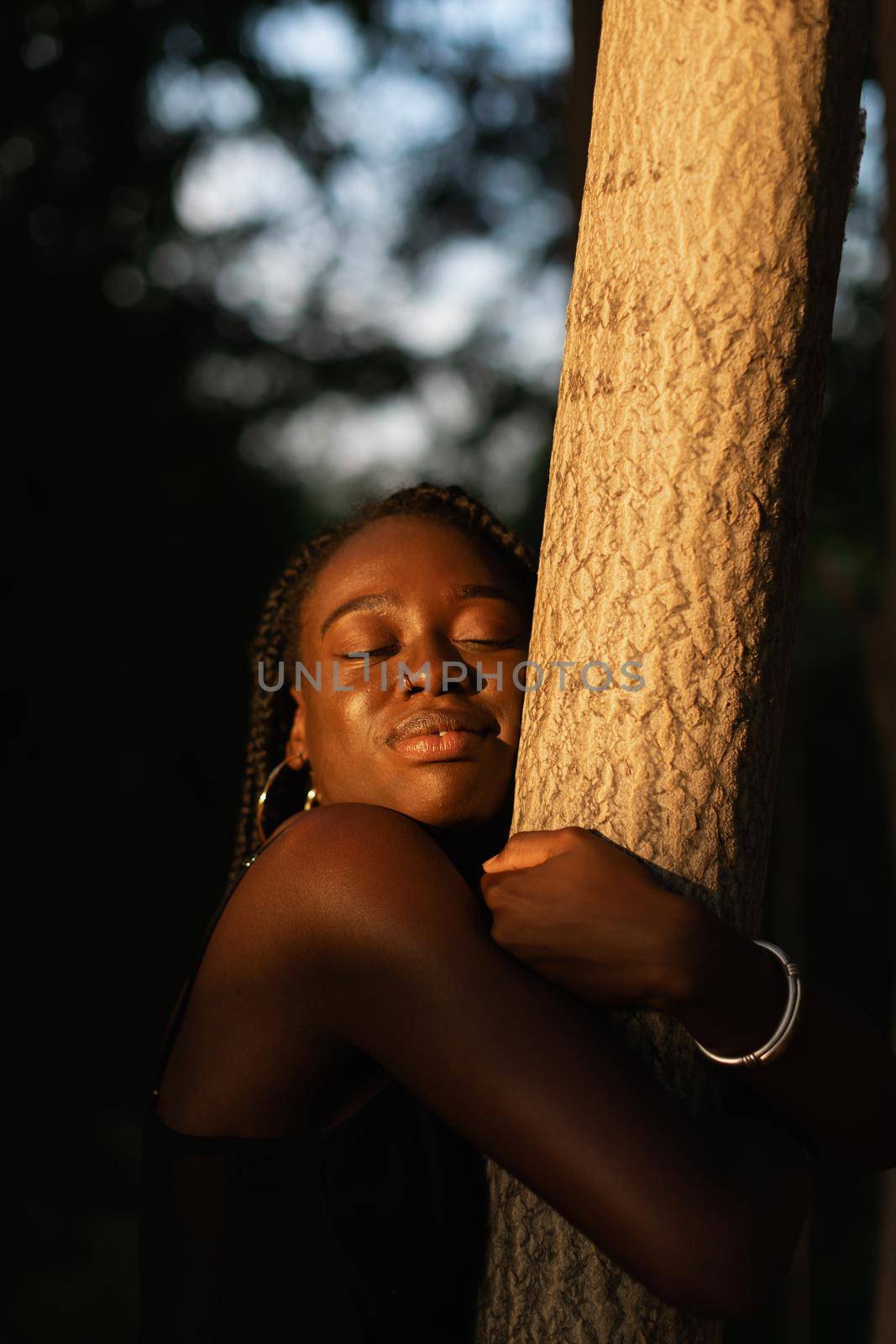 Close-up portrait of a young black woman embracing a tree with love while closing her eyes at the park during sunset