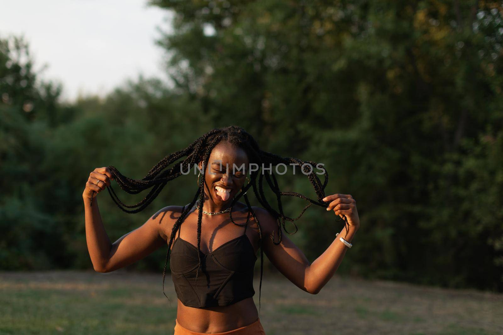 Portrait of a young black female playfuly moving her braids while sticking out her tongue at the park by stockrojoverdeyazul