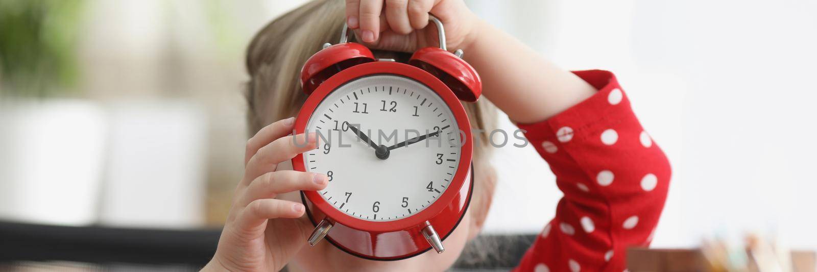 Little girl holding red alarm clock in front of her face. Sleep and wakefulness in children concept