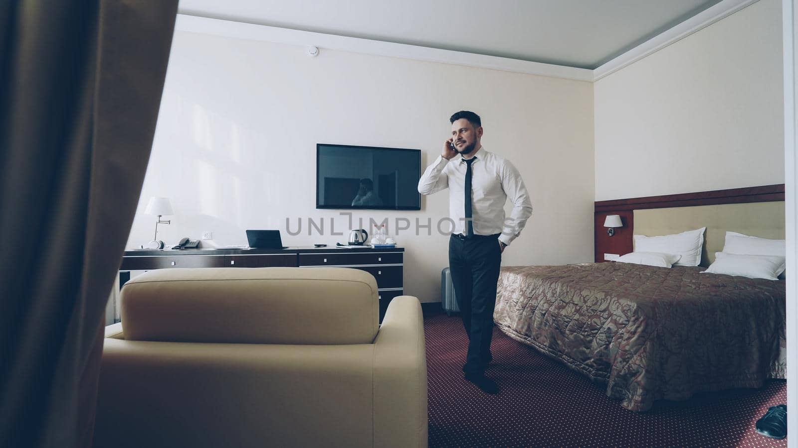 Confident businessman talking on mobile phone while walking around hotel room. Travel, business and people concept by silverkblack