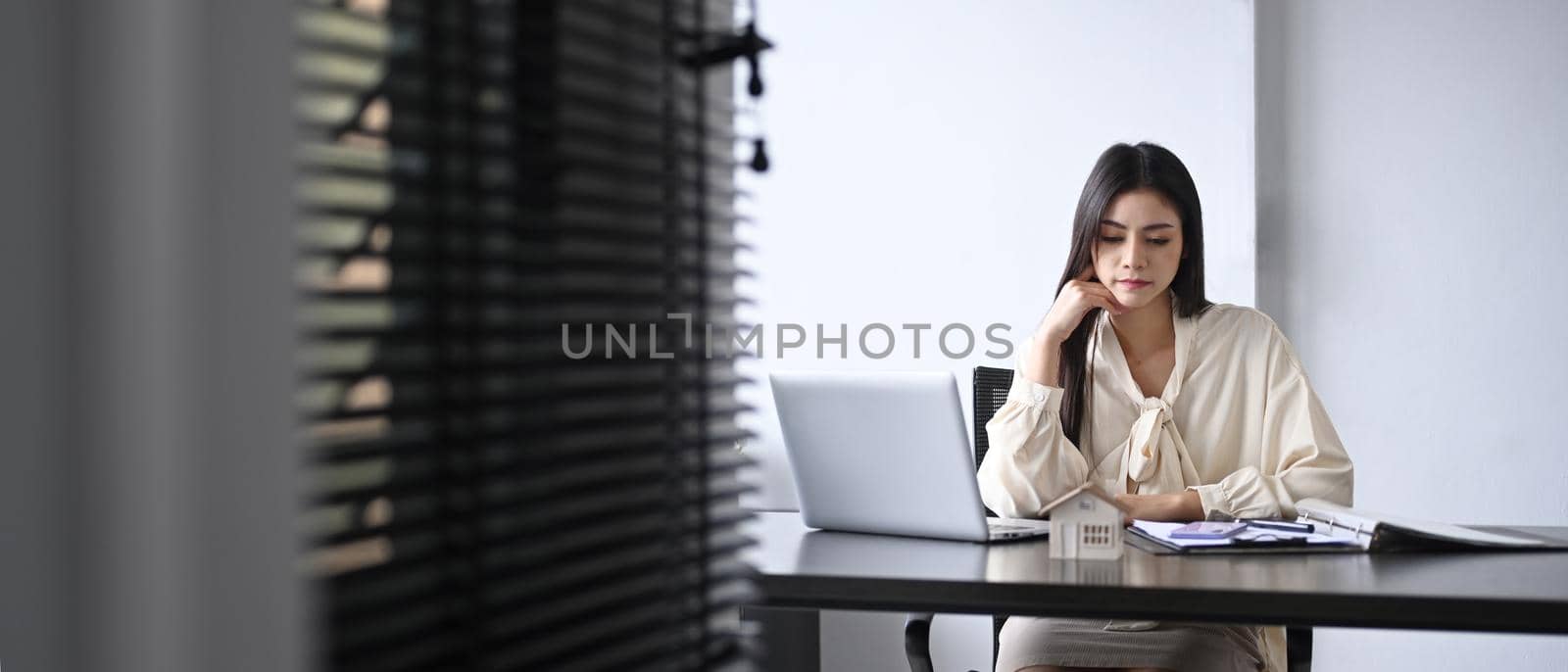 Pensive young businesswoman working with document and laptop while sitting in her personal office.