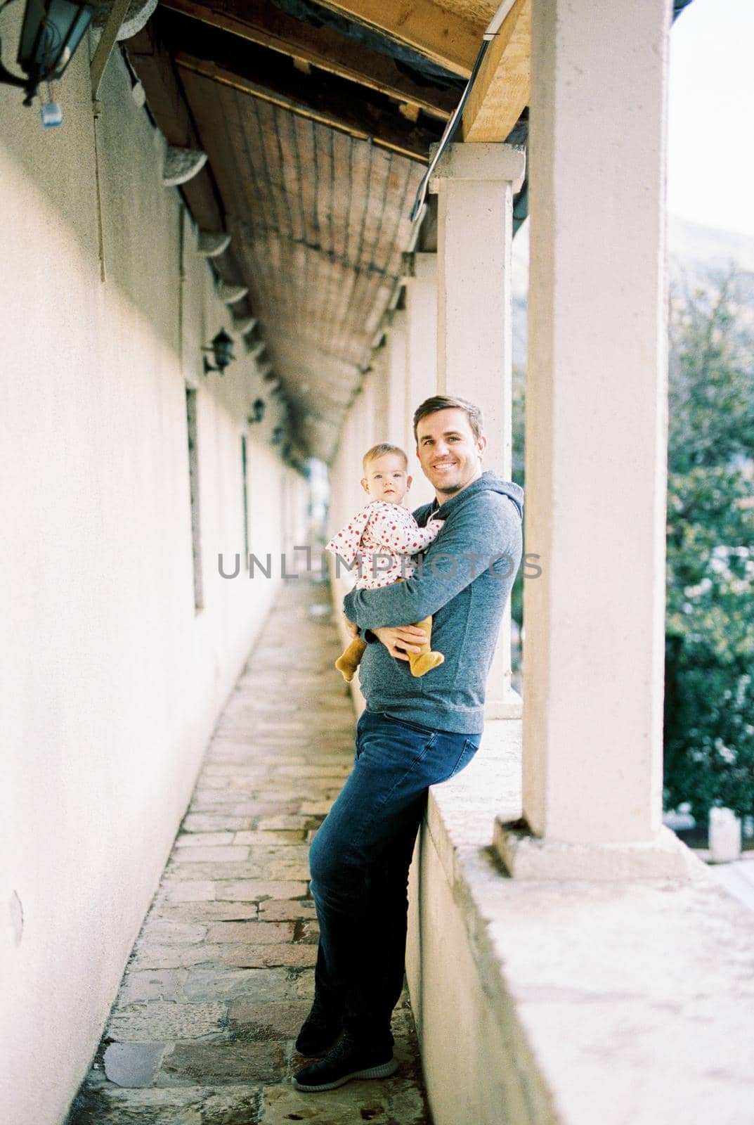 Smiling dad with a baby in his arms stands on the terrace leaning against a stone fence by Nadtochiy