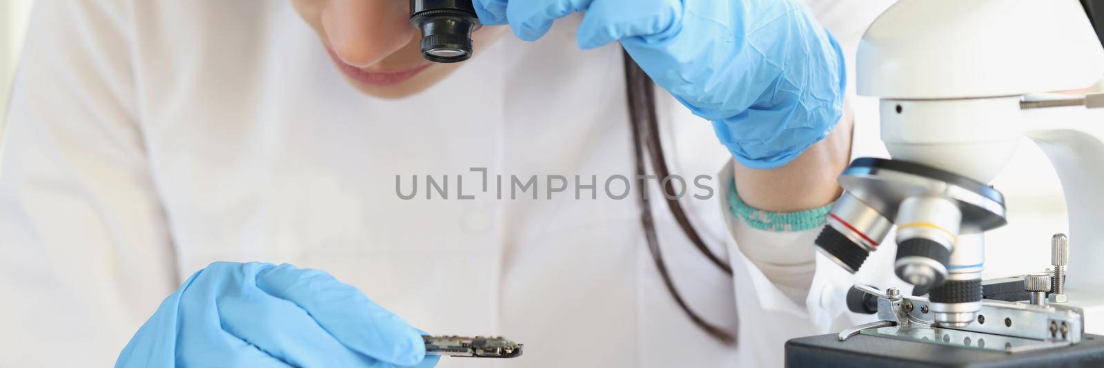 Woman scientist looking through magnifying glass at computer chip near microscope in laboratory by kuprevich