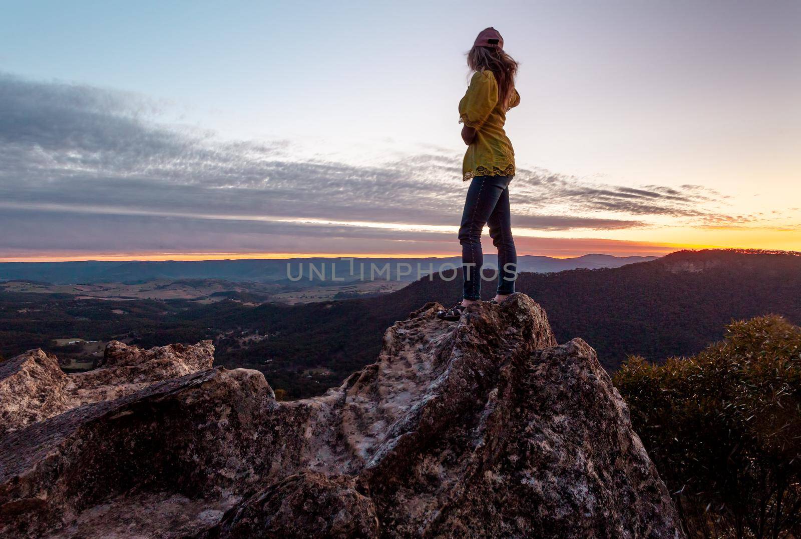 A casually dressed woman stands high on a rock with her hair and top blowing with the brisk wind, her view  overlooks the mountains and valleys and she is watching the sun set.
