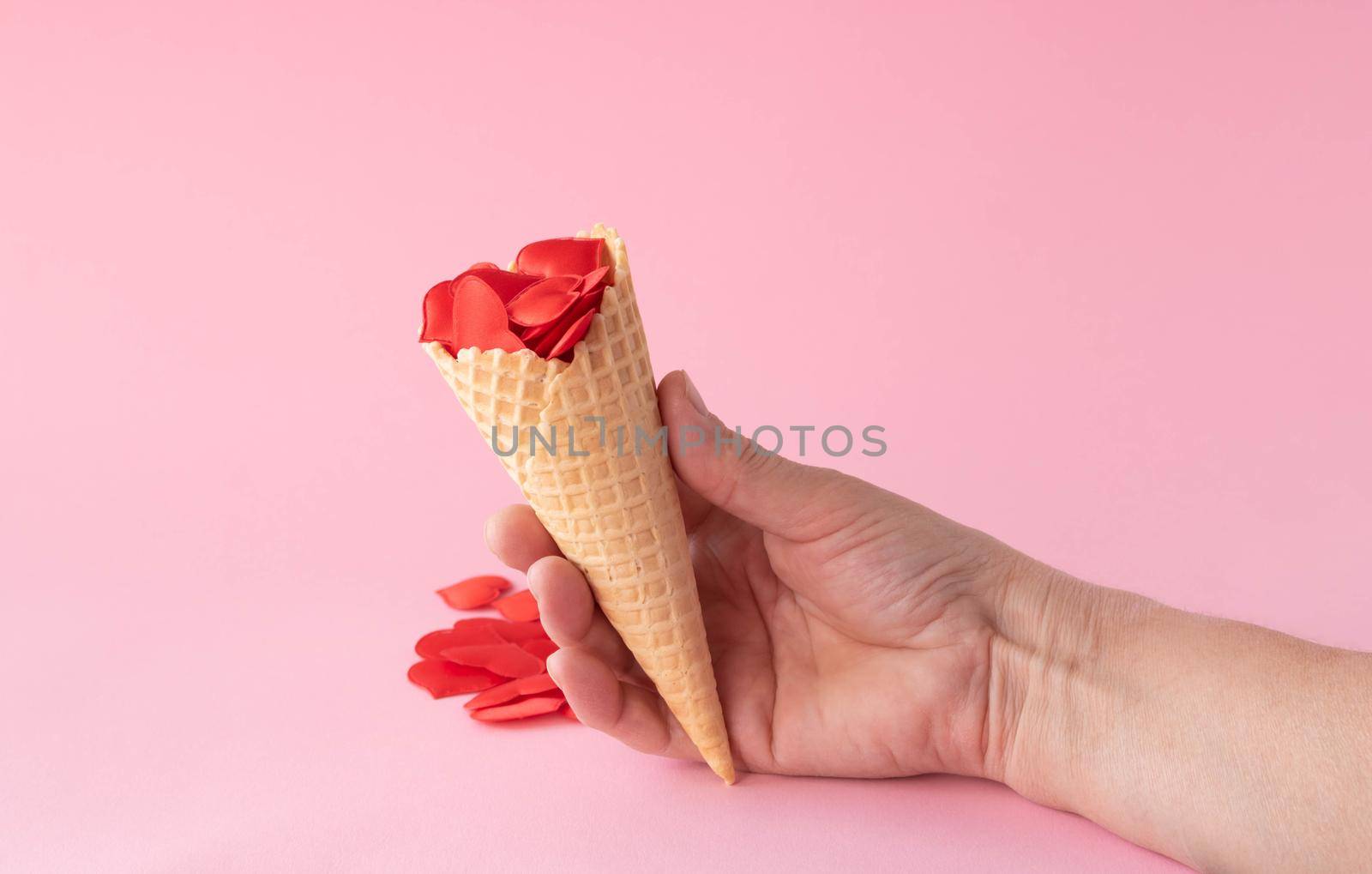 The hand holds an ice cream cone with red hearts. Creative photo of empty waffle cones with scattered red hearts on a pink background. The concept of love, summer, spring.