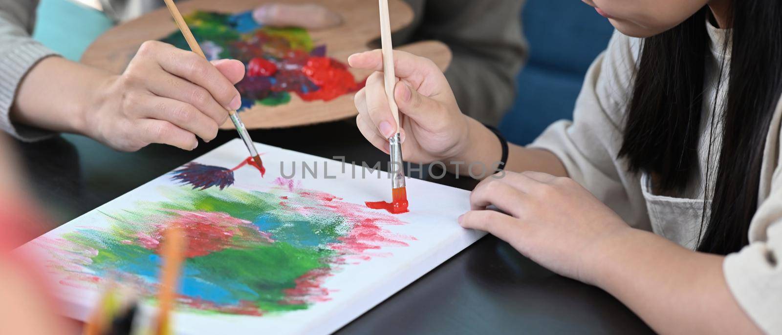 Cropped shot little girl painting picture in art class with teacher. by prathanchorruangsak