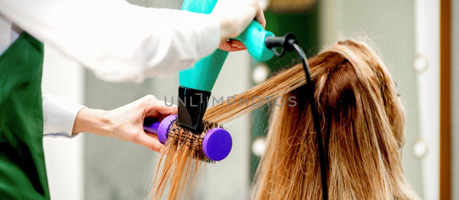 Back view of young woman receiving drying hair with a hairdryer and hairbrush in a hair salon