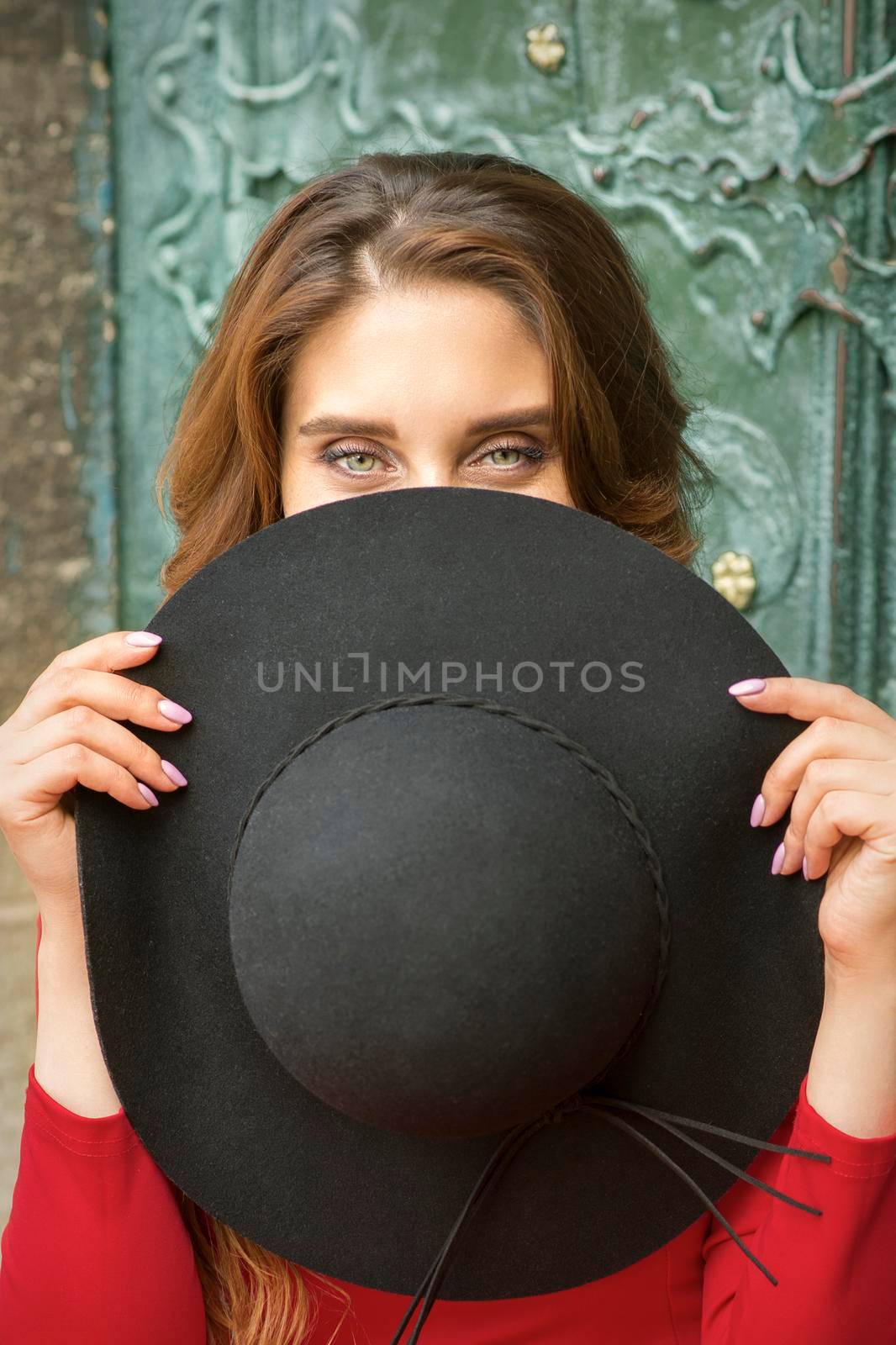 A fashionable beautiful young caucasian woman hiding face with a round black hat on the background the ancient door