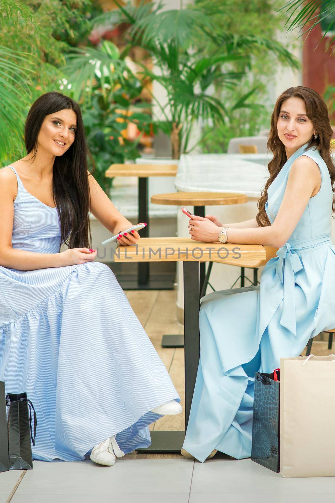 Two beautiful young women sitting at the table with smartphones and shopping bags looking at camera in a cafe outdoors