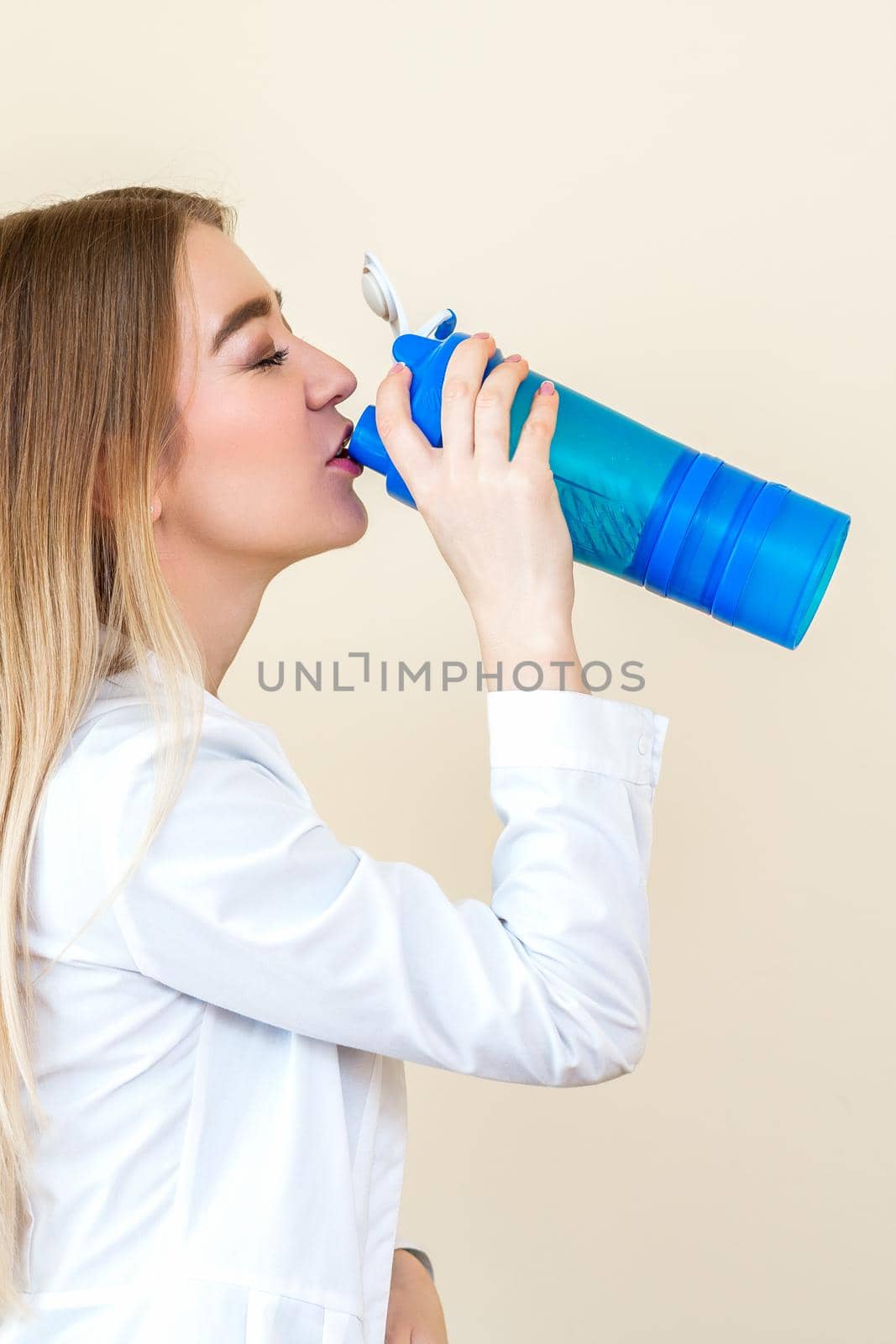 Side view of beautiful young caucasian woman is drinking water from plastic bottle against a beige background