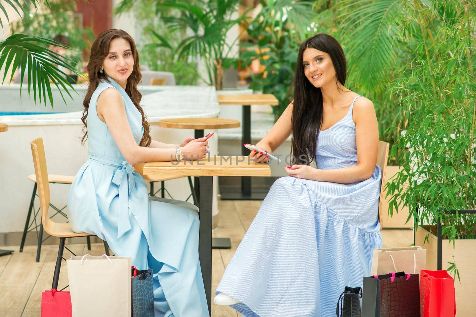 Two beautiful young women sitting at the table with smartphones and shopping bags looking at camera in a cafe outdoors